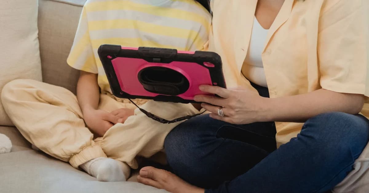 An adult and a child sit close together on a pale sofa, looking at a tablet in a pink case.